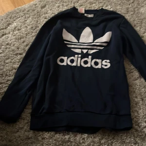 Svart Adidas sweatshirt med stor logga - Säljer en svart långärmad sweatshirt från Adidas med stor vit trefoil-logga och text på bröstet. Tröjan har klassisk passform och är gjord i mjuk bomullsmix, perfekt för en sportig och avslappnad stil.