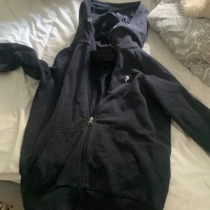 Svart hoodie från Peak Performance - Mysig svart hoodie från Peak Performance med dragkedja framtill och stor huva. Klassisk design med fickor på magen och diskret logga på bröstet. Perfekt för chill dagar eller när du vill ha något bekvämt och stilrent.