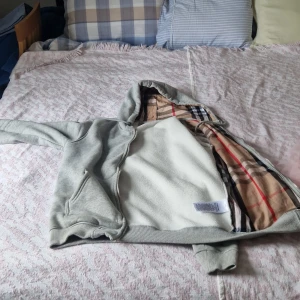 Burberry kofta - ANVÄND 1-2 GÅNGER. FÖR LITEN FÖR MIG. Snygg grå hoodiejacka från Burberry med dragkedja och klassiskt rutigt foder i huvan. Jackan har två fickor framtill, ribbade muddar och snörning i huvan. Perfekt för dig som vill ha en stilren och bekväm look med en touch av lyx.