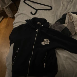 Svart Moncler hoodie med dragkedja - Svart hoodie från Moncler med huva och hel dragkedja framtill. Tröjan har två fickor med dragkedjor och en snygg Moncler-logga på bröstet. Perfekt för dig som gillar stilrena och exklusiva plagg med streetkänsla.