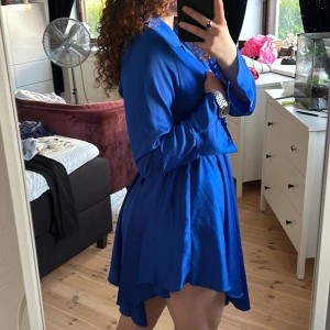 Blå skjortklänning med bälte - Stylish blue shirt dress with a gold chain belt at the waist. The dress has long sleeves, buttons at the front and a slightly flared skirt. Perfect for those who want a stylish and trendy look. Can fit from size XS-M depending on how tall you are!
