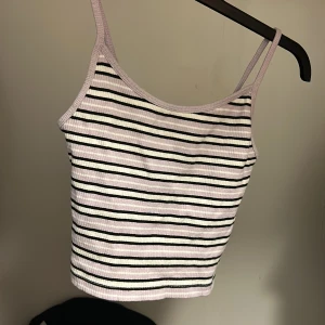 Randig ribbad linnetopp - Säljer en ribbad linnetopp med smala axelband. Toppen är vit med svarta och ljuslila ränder och har en croppad passform. Perfekt för varma dagar eller som ett lager under en skjorta.