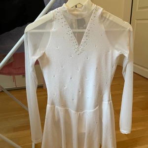 Vit konståkningsklänning med kristaller från Sagester - Superfin vit konståkningsklänning från Sagester med meshärmar och kristalldetaljer framtill och baktill. Klänningen har v-ringning med kristaller och en snygg öppning i ryggen. Perfekt för isdans eller liknande aktiviteter. Den är köpt för 1460 kr. Jag har haft den under en säsong och är därför fortfarande mycket bra i skick.