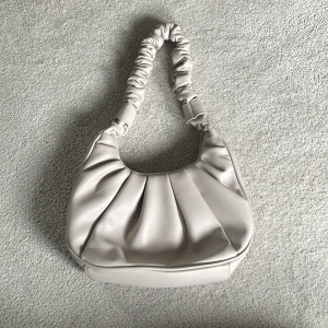 Beige axelväska med rynkad rem - Säljer en beige axelväska i syntetmaterial med snygga veck framtill och en rynkad axelrem. Säljer då den aldrig är använd🤍