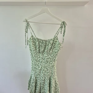 Ljusgrön blommig jumsuit med knytband - Supersöt ljusgrön jumpsuit(klänning) med små svarta blommor och smala axelband som knyts. Klänningen har en lätt utställd kjol och smockad överdel för en bekväm passform. Perfekt för varma dagar och somriga tillfällen.