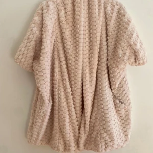 Beige teddyjacka från KS.selection stl 38 - Mysig och oversized beige teddyjacka från KS.selection med korta ärmar och ett unikt rutigt strukturmönster. Jackan är tillverkad i mjuk polyester och har en avslappnad passform, perfekt för lager-på-lager-stil. Passar både som jacka/kavaj inne vid fest eller utomhus under vår och sommar. Perfekt för att lyfta en outfit lite extra! 