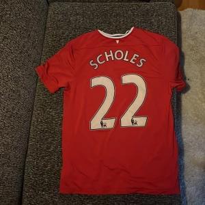 Retro Manchester United fotbollströja - Röd Manchester United matchtröja med Scholes och nummer 22 på ryggen. Tröjan har korta ärmar, vitt tryck och klubbmärke på bröstet. Nike-logga och Aon sponsortryck på framsidan. Klassisk vit och svart krage. Retro och är äkta 