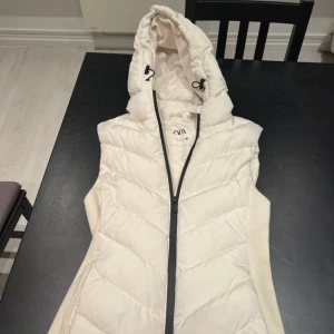 Vit dunväst med huva från Zara - Snygg vit dunväst från Zara med dragkedja framtill och stor huva med dragsko. Västen har ett quiltat mönster och är perfekt för lager-på-lager-stil. Passar till många olika outfits och håller dig varm under kyliga dagar.