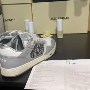Grå och vita Dior sneakers med monogram - Säljer ett par grå och vita Dior sneakers med klassiskt Dior-monogram på sidan. Skorna har snörning, rund tå och platt sula. Perfekta för dig som vill ha en lyxig och stilren look. Äkta finns kvitto och allting box är borta men har kvar kvittot! Priset kan diskuteras måttligt