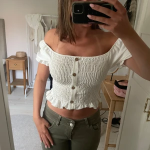 Vit topp med knappar - Säljer en söt vit croppad blus med smockat tyg och volangkant nertill. Blusen har korta puffärmar och knappar framtill. Perfekt för varma dagar och ger en romantisk känsla.