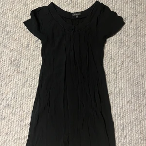 Svart kortärmad klänning - En enkel svart klänning med korta ärmar och rund halsringning. Klänningen har en rak passform och är tillverkad i ett lätt, något skrynkligt material. Perfekt för dig som gillar stilrena och minimalistiska plagg.