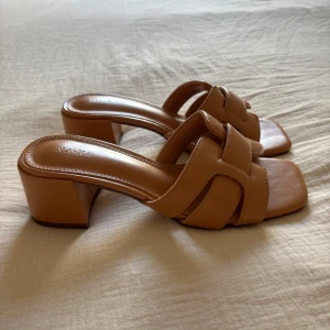 Bruna klackade sandaler från NA-KD - Snygga bruna sandaler med klack från NA-KD. Skorna har en fyrkantig tå, breda flätade remmar och öppen häl. Perfekta för varmare dagar och ger en trendig look till din outfit. Aldrig använda!