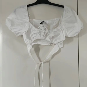 Vit croppad blus med puffärmar från H&M  - Säljer en vit croppad blus från H&M med puffiga korta ärmar och knytband i midjan. Blusen har en söt, romantisk stil med resår vid halsringningen och under bysten. Perfekt för sommardagar!