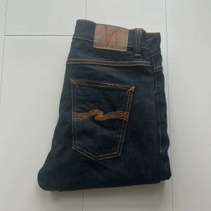 Mörkblå jeans från Nudie Jeans Lean Dean nya utan tags - Säljer ett par mörkblå jeans från Nudie Jeans med klassiska orange sömmar och logga på bakfickan. Jeansen har rak passform och är tillverkade i denim med snygga detaljer. Perfekta för dig som gillar stilrena och tidlösa jeans.