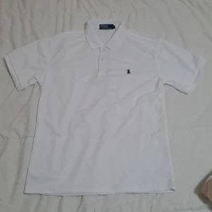 Vit pikétröja från Polo Ralph Lauren - Klassisk vit pikétröja från Polo Ralph Lauren med broderad logga på bröstet. Tröjan har krage, knappar framtill och korta ärmar. Perfekt för en stilren och sportig look.