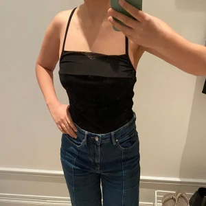 Svart croppad linnetopp med smala axelband - Säljer en svart croppad linnetopp med smala axelband som korsas i ryggen. Toppen har en rak urringning och sitter tight mot kroppen. Perfekt att styla med jeans eller kjol.