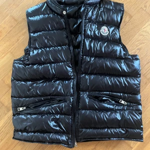 Svart dunväst från Moncler - Säljer en glansig svart dunväst från Moncler med två dragkedjefickor framtill och Moncler-logga på bröstet. Västen har hög krage och stängs med dragkedja och tryckknapp upptill. Perfekt för lager-på-lager under kyliga dagar.
