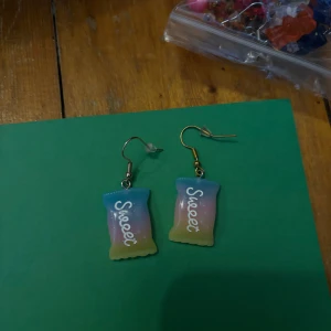 Färgglada örhängen med texten 'Sweet' - Unika örhängen i form av små påsar med texten 'Sweet'. De har en färgskiftning i blått, rosa och gult och är perfekta för dig som gillar lekfulla accessoarer.