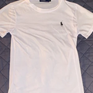 Vit t-shirt från Polo Ralph Lauren - Klassisk vit t-shirt från Polo Ralph Lauren med rund halsringning och liten broderad logga på bröstet. T-shirten har korta ärmar och är tillverkad i mjuk bomull. Perfekt basplagg till garderoben.