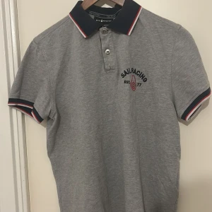 Grå piké från Sail Racing - Snygg grå pikétröja från Sail Racing med mörkblå krage och ärmslut, samt röda och vita detaljer. Broderad logga på bröstet och liten etikett på sidan. Perfekt för en sportig och stilren look.