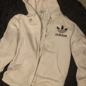 Vit hoodie från Adidas med dragkedja - Säljer en vit hoodie från Adidas med dragkedja framtill och två fickor. Tröjan har Adidas klassiska logga i svart på bröstet och huva med dragsko. Perfekt för en sportig och avslappnad stil.