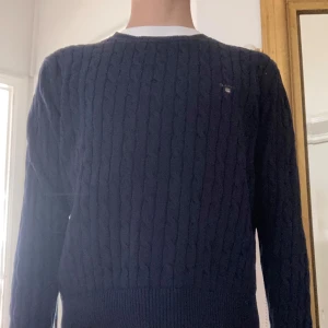 Blå kabelstickad tröja från Gant - Säljer en klassisk blå kabelstickad tröja från Gant med rund halsringning. Tröjan har långa ärmar och ett broderat Gant-logo på bröstet. Perfekt för en stilren och tidlös look.