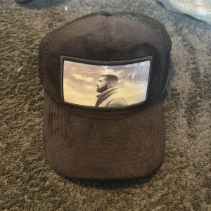 Svart keps från Icons Living med tryck - Svart keps med mesh på sidorna och justerbart spänne bak. Framtill finns ett tryck med ett porträttmotiv i svartvitt. Märkt med 'ICONS LIVING' på insidan. Perfekt för en cool och avslappnad stil.