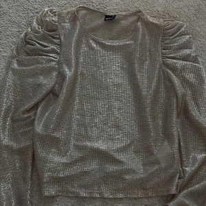 Glittrig silver topp med puffärmar från Gina Tricot - Säljer en glittrig silverfärgad topp från Gina Tricot med långa puffärmar och rund halsringning. Toppen har en figurnära passform och är perfekt för att sticka ut på festen.