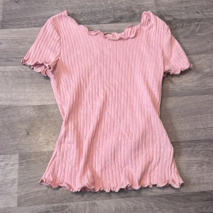 Rosa ribbad topp från SHEIN - Säljer en ljusrosa ribbad topp från SHEIN med volangdetaljer vid halsringning, ärmslut och nederkant. Toppen har korta ärmar och är tillverkad i mjuk bomullsblandning. Perfekt för en söt och avslappnad look!