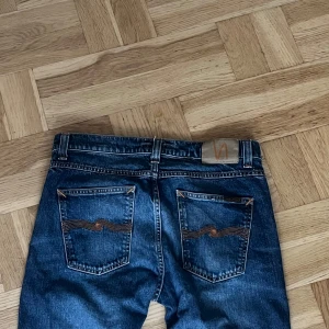 Mörkblå jeans från Nudie Jeans - Säljer ett par klassiska mörkblå jeans från Nudie Jeans med raka ben och snygga detaljer på bakfickorna. Jeansen har normal passform och är tillverkade i slitstarkt denim. Perfekta för dig som gillar stilrena och tidlösa jeans. Modell lean Dean 31/32