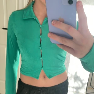 Grön croppad topp med knappar - Snygg grön croppad topp med långa ärmar och krage. Toppen har dekorativa knappar framtill och är i ett mjukt, stretchigt material. Perfekt att styla till både jeans och kjol. Lappen är kvar 