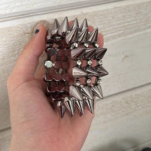 Silvrigt armband med nitar och strasstenar - Coolt armband i silverfärgad metall med stora konformade nitar och glittrande strasstenar mellan nitarna. Armbandet är elastiskt och passar de flesta handleder. Perfekt för dig som vill sticka ut med en edgy accessoar.
