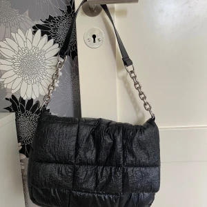 Svart quiltad axelväska från HM Divided - Säljer en svart quiltad axelväska från Divided med kedjedetaljer. Väskan har en puffig design och stängs med tryckknapp. Perfekt för att bära dina viktigaste saker med stil.
