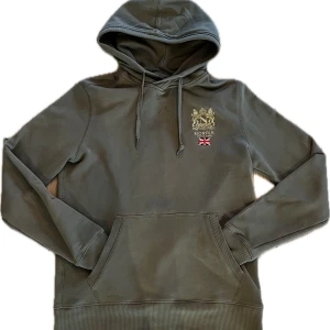 Olivgrön hoodie från Morris i storlek S  - Säljer en olivgrön hoodie från Morris med broderad logga och brittisk flagga på bröstet. Tröjan har huva med dragsko och en stor magficka. Perfekt för dig som gillar stilrena detaljer och bekväma plagg. Även i ett gott skick!