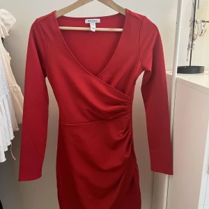 Röd omlottklänning från NLY ONE - Snygg röd omlottklänning från NLY ONE i figurnära modell. Klänningen har lång ärm, v-ringning och draperad detalj i midjan. Stängs med dragkedja i sidan. Perfekt för dig som vill sticka ut med en klassisk look.