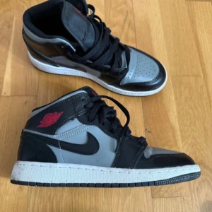 Nike Air Jordan 1 Mid svart och grå - Säljer ett par Nike Air Jordan 1 Mid i svart och grå med röda detaljer. Klassisk design med svart swoosh, perforerad tå och högt skaft. Skorna har snörning och vit sula. Perfekta för dig som gillar streetwear och sneakers.