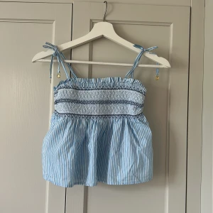 Blå- och vitrandig linnetopp med smock - Söt blå- och vitrandig linnetopp med smock upptill och tunna axelband som knyts. Toppen har volangkant vid bysten och är luftig nertill. Perfekt för varma dagar.