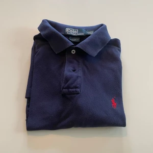 Mörkblå pikétröja från Polo Ralph Lauren - Säljer en klassisk mörkblå pikétröja från Polo Ralph Lauren med röd broderad logga på bröstet. Tröjan har krage, knappar vid halsen och är kortärmad. Perfekt för en stilren och avslappnad look. Storlek L