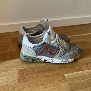 Grå och vita sneakers med orange detalj - Säljer ett par grå och vita sneakers från Premiata med orange detalj på sidan och vit snörning. Skorna har en platt sula med tryck och rund tå. Perfekta för dig som gillar en sportig och trendig stil.