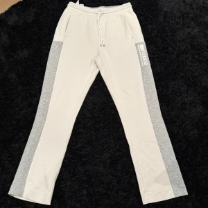 Vita raka mjukisbyxor med gråa sidor från BOSS - Snygga vita mjukisbyxor från BOSS med gråa paneler längs sidorna och logga tryckt på vänster ben. Byxorna har dragsko i midjan och sidofickor. Perfekta för en avslappnad stil.