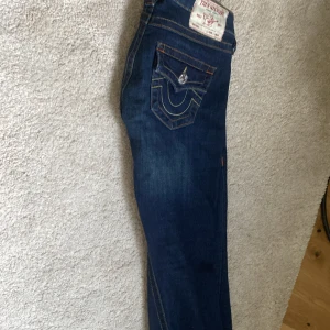 Mörkblå jeans från True Religion - Säljer ett par mörkblå jeans från True Religion med klassisk kontrastsöm och snygga detaljer på bakfickan. Byxorna har normal passform och raka ben. Perfekta för dig som gillar en tidlös jeansstil. Dom är boocut. Egentligen är dom i storlek 23, men jag har gjort dom ungefär 4 cm kortare för jag är ganska kort. Så jag skulle säga att dom passar någon som är kring 155-163 cm lång❤️Jag har använt dom 1 gång så dom är som helt nya. Jag säljer dom för att jag vill ha dom i en ljusare färg!