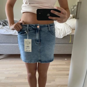 Blå jeanskjol från Miss Bon - Säljer en klassisk blå jeanskjol från Miss Bon. Kjolen har fem fickor, knapp och dragkedja framtill samt bälteshällor. Perfekt basplagg med en snygg, enkel design och kort längd.