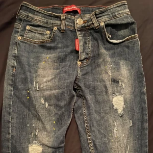 Dsquared2 slitna jeans med färgstänk - Mörkblå jeans från Dsquared2 med slitningar, små gula färgstänk och klassisk femficksdesign. Byxorna har en gul patch med logga bak och röda Dsquared2-detaljer vid gylfen. Perfekt för dig som gillar en edgy och trendig look.