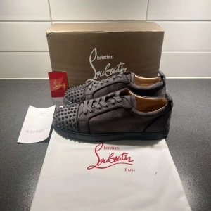 Grå mocka sneakers med nitar från Christian Louboutin - Säljer ett par grå sneakers från Christian Louboutin i mocka med coola nitar på tån. Skorna har rund tå, platt sula och snörning. Perfekta för dig som vill sticka ut med en lyxig och edgy stil.