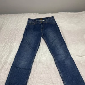 Blå jeansbyxor från Lee - Säljer ett par klassiska blå jeans från Lee med raka ben och normal passform. Byxorna har fem fickor, gylf med dragkedja och knapp samt Lee-logga på bakfickan och bakre linningen. Perfekta till vardags och enkla att matcha med olika stilar. Storlek 12-13 år 160 cm.