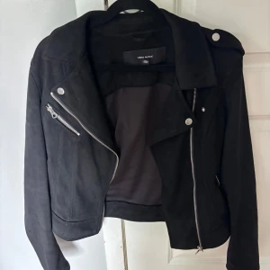 Svart moccajacka från Vero Moda - Säljer en svart moccajacka från Vero Moda med silvriga dragkedjor och knappar. Jackan har klassisk krage, snedställd dragkedja och detaljer på axlarna. Perfekt för en cool och stilren look. Väldigt tunn och skön mocka