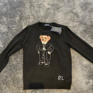 Svart stickad tröja med björn från Polo Ralph Lauren - Svart stickad tröja från Polo Ralph Lauren med en ikonisk björn i kostym på bröstet och RL-broderi nedtill. Rund halsringning och ribbade muddar vid ärmslut och nederkant. Perfekt för dig som gillar klassisk stil med en twist. Helt oanvänd, priset är diskuterbart.