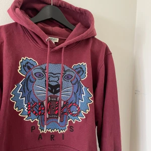Vinröd Kenzo hoodie med tigerbrodyr - Säljer en vinröd hoodie från Kenzo med ikonisk tigerbrodyr och logga framtill. Tröjan har huva med snörning och är tillverkad i mjuk bomull. Perfekt för dig som vill sticka ut med en fet streetstyle-look. Nypris: 4299kr