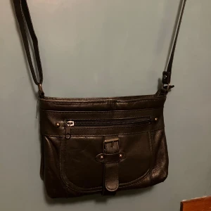 Svart axelväska i skinn med dragkedja - Snygg svart axelväska i skinn med justerbar rem, flera fack med dragkedja både fram och bak samt ett spänne som detalj på framsidan. Perfekt storlek för att få plats med allt viktigt och enkel att matcha till olika outfits.