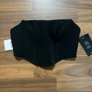 Svart korsett-topp NA-KD x Moa Mattsson - Snygg svart korsett-topp från NA-KD x Moa Mattsson. Toppen är axelbandslös med markerade sömmar och en stilren, croppad passform. Perfekt att styla med höga jeans eller kjol för en trendig look.